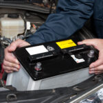 Mechanic in a blue uniform holding and removing a car battery from the engine bay.