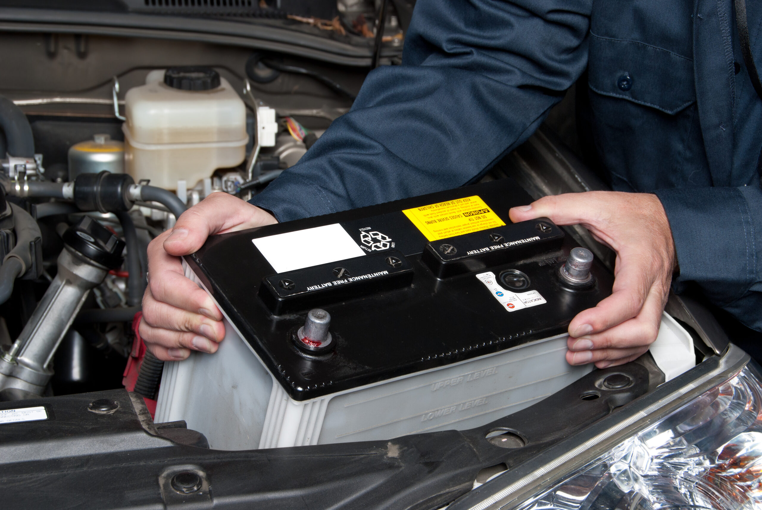 Mechanic in a blue uniform holding and removing a car battery from the engine bay.