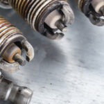 Used spark plugs with carbon buildup next to a clean new spark plug, illustrating signs of wear and tear that can affect engine performance.