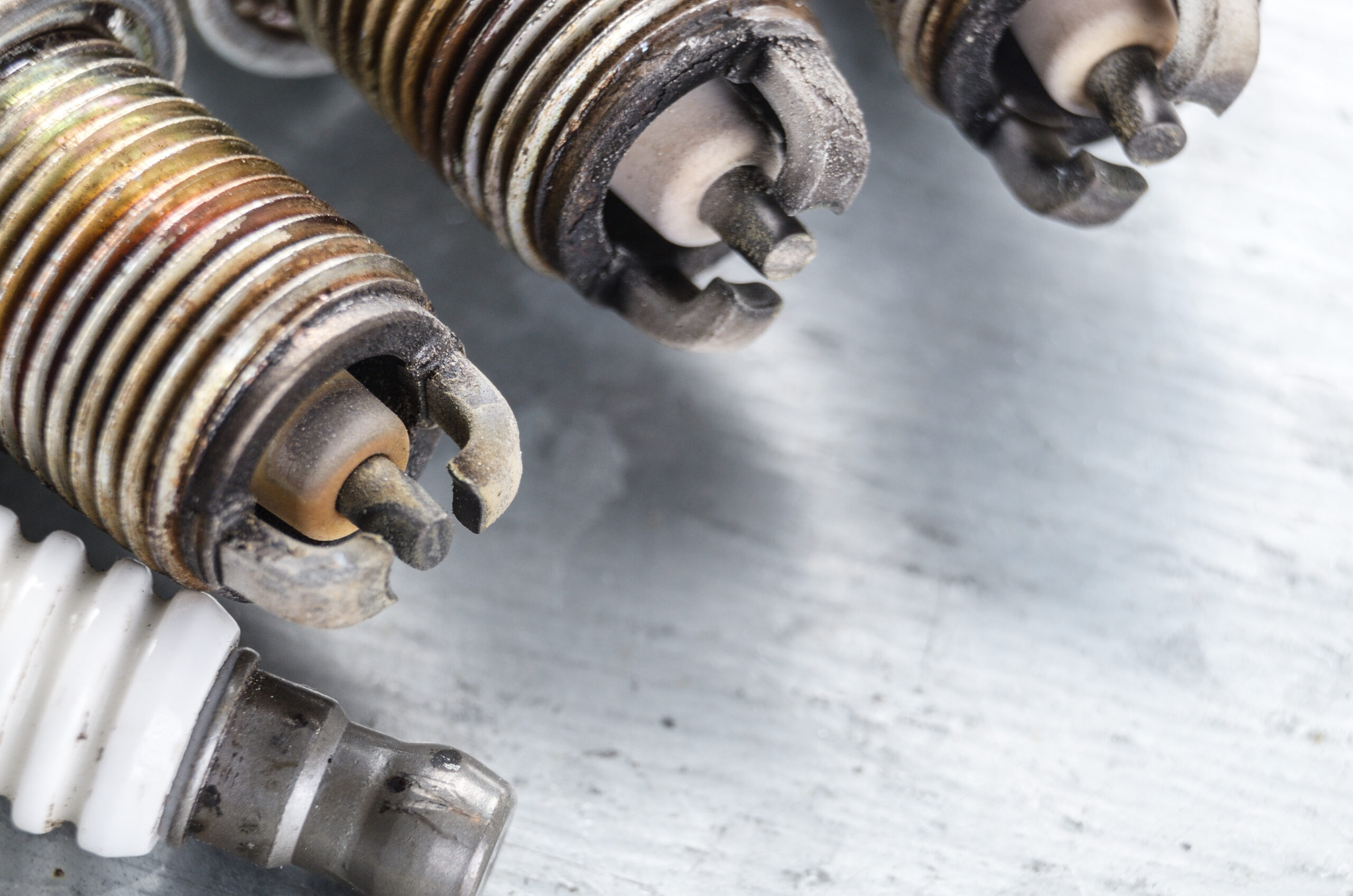 Used spark plugs with carbon buildup next to a clean new spark plug, illustrating signs of wear and tear that can affect engine performance.